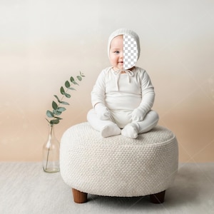 Op de afbeelding: Een baby in een witte outfit en muts zit op een witte, ronde poef met houten poten. Links staat een heldere glazen vaas met groene bladeren. De achtergrond is een zachte gradiënt van beige en perzik.