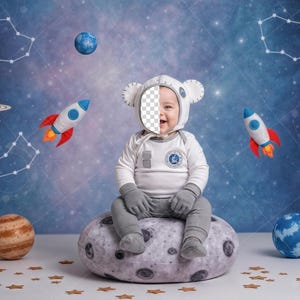以下が含まれることがあります： 白い宇宙飛行士のコスチュームを着た赤ちゃんが、お揃いの帽子と手袋をして、月の形をしたクッションに座っています。背景には、惑星、ロケット、星座が描かれた星空の宇宙シーンがあります。