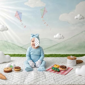 May include: A baby in a pale blue outfit and hat with a bow sits on a white quilted blanket. Kites fly in the sky above a green landscape. Decorative food items and cloud-shaped pillows are arranged on the blanket.