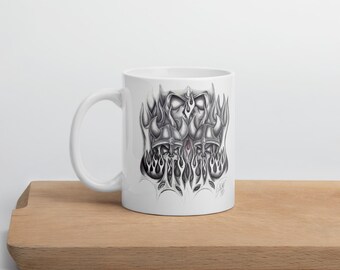 May include: White ceramic mug with a black and grey illustration of a stylized design with flames and helmet shapes. The mug has a curved handle and is on a wooden surface.