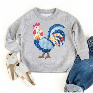 Peut inclure: Un sweat-shirt gris clair avec un motif de coq coloré. Le coq a une crête rose, des détails floraux et un motif à pois et à carreaux bleus. L'image montre également une paire de pantoufles grises et un jean bleu.