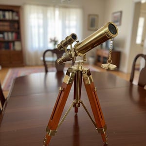 Könnte beinhalten: Ein Messing-Teleskop auf einem Holzstativ. Das Teleskop hat einen langen, zylindrischen Tubus und ein kleineres Sekundärteleskop. Die Stativbeine sind aus Holz mit Messingbeschlägen. Ein dekorativer Artikel.