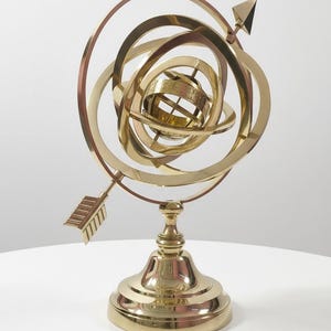 May include: A brass armillary sphere with multiple concentric rings and a central globe. An arrow points through the rings. The sphere sits on a decorative, tiered base. The piece is made of polished brass.