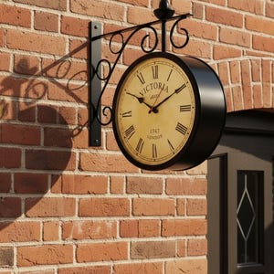 May include: A black, double-sided outdoor clock with the text "VICTORIA HALL 1747 LONDON" on a cream-colored face. The clock has black Roman numerals and hands, and is mounted on a decorative black metal bracket against a red brick wall.