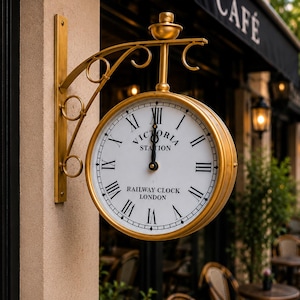May include: A gold-coloured, double-sided wall-mounted clock with a white face, black Roman numerals, and black hands. The clock face features the text "VICTORIA STATION" and "RAILWAY CLOCK LONDON." It is supported by an ornate, curved metal bracket.