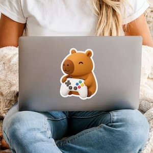 May include: A sticker featuring a cartoon capybara holding a white video game controller with colorful buttons. The capybara is brown with a lighter brown face and a small smile. The sticker is on a gray laptop.