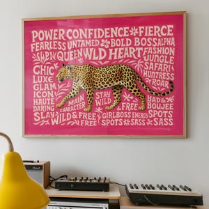May include: A framed pink print featuring a leopard illustration with the words "Power, Confidence, Fierce, Wild" and other empowering words. The leopard is brown with black spots. The frame is a light wood color.
