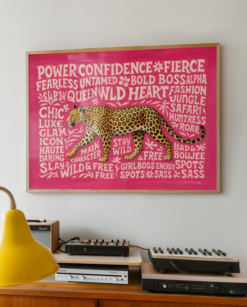 May include: A framed pink print featuring a leopard illustration with the words "Power, Confidence, Fierce, Wild" and other empowering words. The leopard is brown with black spots. The frame is a light wood color.