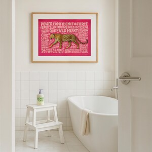 May include: A framed print with a pink background and a leopard illustration. The print includes the words "POWER CONFIDENCE FIERCE" and other empowering words. A white step stool holds a bottle of "good one" soap in a bathroom setting.