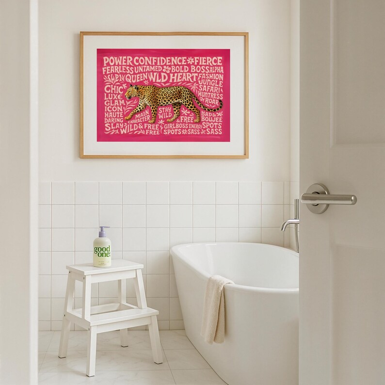 May include: A framed print with a pink background and a leopard illustration. The print includes the words "POWER CONFIDENCE FIERCE" and other empowering words. A white step stool holds a bottle of "good one" soap in a bathroom setting.