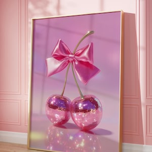 Op de afbeelding: Een ingelijste kunstdruk met twee kersen met discobaloppervlakken en een roze satijnen strik. De kersen zijn verbonden door een gouden steel tegen een roze achtergrond. De lijst is goudkleurig.