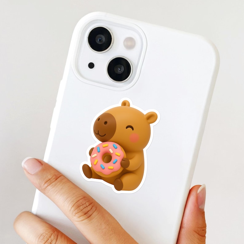 May include: A brown capybara sticker holding a pink donut with blue and white sprinkles. The sticker is on a white phone case. The capybara has a smiling expression.