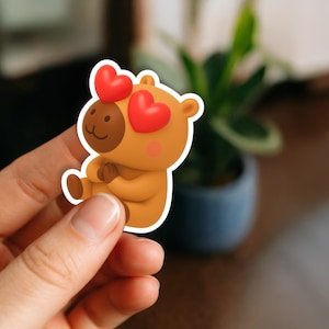May include: A sticker featuring a cartoon bear with red heart-shaped eyes and a brown face. The bear is light brown with a white outline and is holding its paws together. The sticker is held in a person's hand.