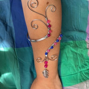 Può includere: Bracciale argentato con un design decorativo in filo metallico. Il bracciale presenta perline rosa e blu e un ciondolo a forma di piuma argentata. Il design include elementi di filo a spirale.