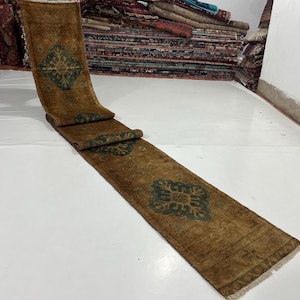 May include: A long, rectangular runner rug with a brown and green color scheme. The rug features a repeating geometric pattern in green. The rug is rolled up in sections, showcasing its length and design. The rug has a fringed edge.