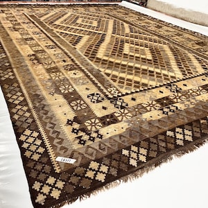 May include: A large, rectangular rug with a geometric pattern in shades of brown, beige, and tan. The rug features a central diamond design and borders with repeating shapes. The rug is displayed on a white surface.