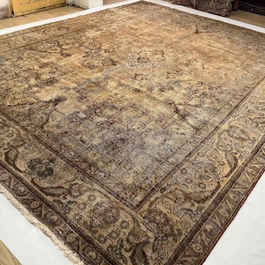 May include: A large rectangular area rug featuring a distressed, faded brown and tan color palette. The rug displays an intricate, symmetrical floral and vine pattern throughout the center field, enclosed by a wide, detailed decorative border.