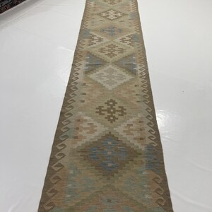 May include: A long, rectangular runner rug with a geometric pattern in shades of beige, brown, and blue. The rug features diamond and cross-like motifs. The edges have a fringe detail. This rug is suitable for hallways or entryways.