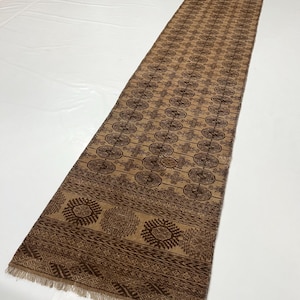 May include: A long, narrow runner rug featuring a tan background with repeating dark brown geometric patterns. The design includes rows of circular motifs and intricate border details, finished with short, light-colored fringe at the ends.