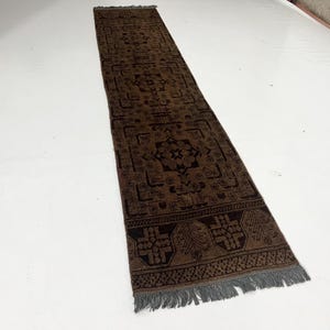 May include: A rectangular brown runner rug with a detailed, dark brown pattern. The rug features a central design with floral and geometric motifs, bordered by a decorative edge and finished with fringe.
