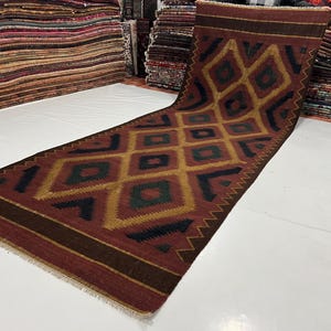 May include: A long, rectangular rug with a geometric diamond pattern in shades of red, gold, green, and navy blue. The rug has a dark brown border and fringe on the ends. The rug is displayed on a white surface.