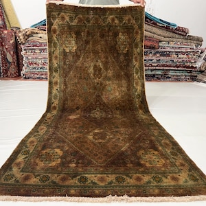May include: A brown and green patterned rug with a detailed floral design. The rug has a rectangular shape with a fringe on the bottom edge. The rug is displayed in a room with other rugs in the background.