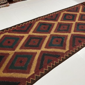 May include: A rectangular rug with a geometric pattern. The rug features a repeating diamond design in shades of red, gold, and teal, with a dark brown border. The rug is likely made of woven fabric.