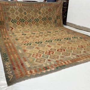 May include: A large rectangular area rug featuring a repeating diamond pattern in shades of tan, beige, green, and rust orange. The rug has a wide decorative border with geometric motifs and a fringed edge along the bottom horizontal side.