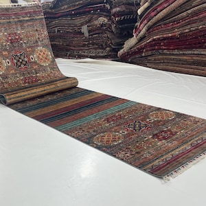 May include: A rolled-out, rectangular rug with a detailed, patterned design. The rug features a mix of colors, including brown, red, and blue, with a striped pattern on the bottom half. The rug is displayed on a white surface.