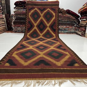 May include: A long, rectangular rug with a geometric diamond pattern in shades of red, gold, green, and black. The rug has a fringe at the bottom and is displayed in a room with other rugs stacked in the background. The rug's design is symmetrical.