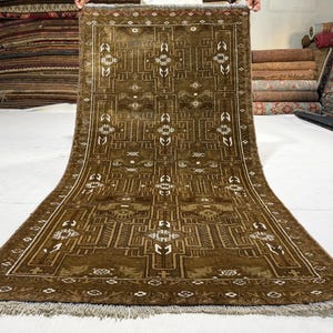 May include: A rectangular area rug featuring a deep olive-brown base color with intricate geometric patterns in white and light brown. The design includes symmetrical linear motifs and a border with small repeating shapes, finished with fringe.