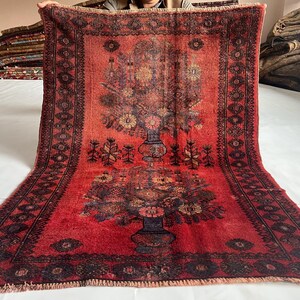 May include: A red and black floral patterned rug. The rug features a central vase design with flowers and foliage. The border has a repeating geometric pattern. The rug is displayed on a white surface.