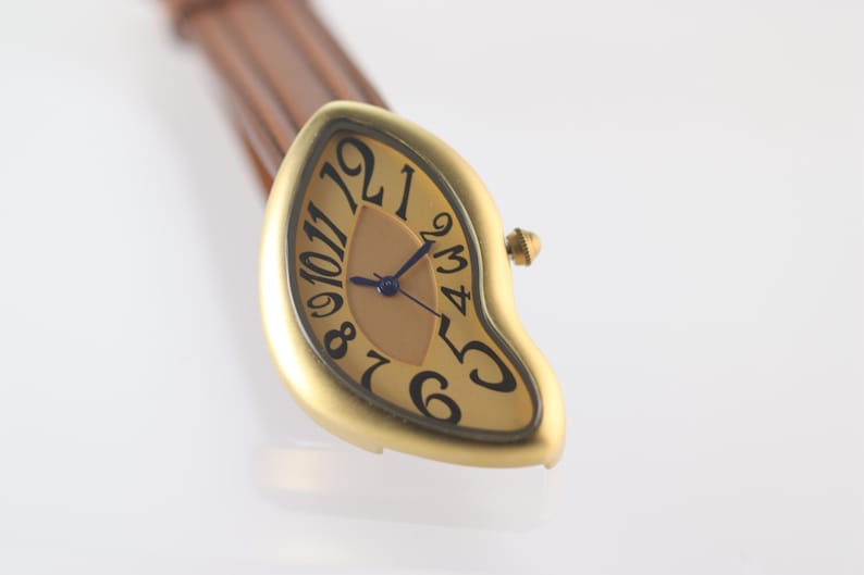 May include: A gold-toned wristwatch with a unique, melted-looking design. The watch face is a light brown color with black Roman numeral hour markers and blue hands. The watch is attached to a brown leather strap.