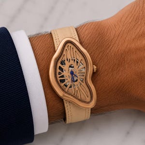 May include: A gold-toned wristwatch with a unique, flowing design. The watch features a cream-colored leather band and a skeletonized face with blue accents. The watch is worn on a wrist, with a navy blue suit jacket visible in the background.