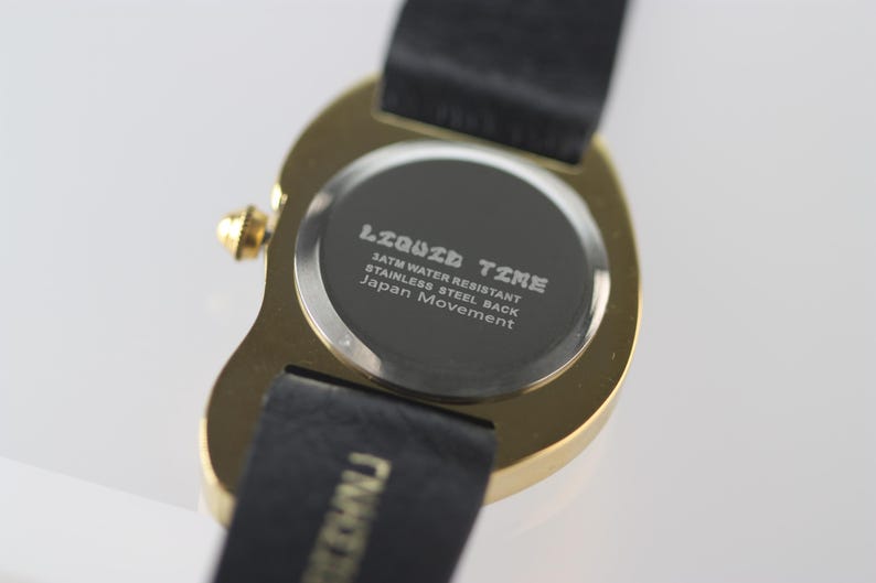 May include: The back of a gold-tone watch with a black leather band. The watch face says "Liquid Time" and "3ATM Water Resistant, Stainless Steel Back, Japan Movement".