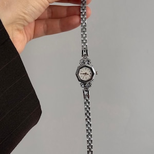 May include: A silver wristwatch with an octagonal face and a linked bracelet. The watch face is white with black Roman numerals and black hands. The bracelet is held by a hand.