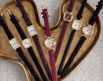 Op de afbeelding: Een verzameling vintage polshorloges met goudkleurige kasten en leren bandjes in verschillende kleuren, waaronder zwart, bordeauxrood en dieprood. De horloges hebben rechthoekige en ovale wijzerplaten met Romeinse cijfers, gepresenteerd op een hartvormige gouden schaal.