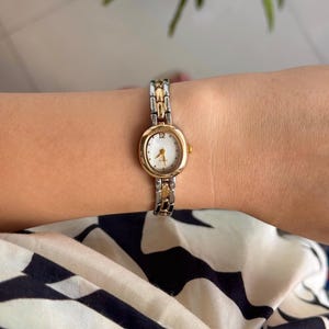 May include: A gold and silver-toned wristwatch with a white face and gold hands. The watch has a small oval face and a linked band. The watch is on a wrist.
