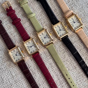 Könnte beinhalten: Eine Auswahl an rechteckigen Armbanduhren mit goldfarbenen Gehäusen und Lederarmbändern in verschiedenen Farben, darunter Burgunderrot, Grün, Schwarz und Beige. Die Zifferblätter haben römische Ziffern und filigrane Zeiger.