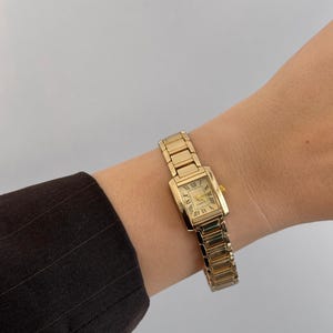 Puede incluir: Un reloj de pulsera rectangular dorado con una correa dorada. La esfera del reloj muestra números romanos y agujas doradas. El reloj está en una muñeca y la correa está hecha de eslabones rectangulares.