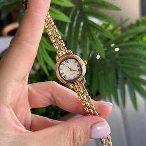May include: A gold-toned wristwatch with a round face and Roman numeral hour markers. The watch has a gold link band and a white watch face. The watch is held up against a background of green foliage.