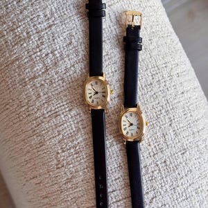 May include: Two gold-toned, oval-faced watches with black leather bands. The watch faces have Roman numerals and a white background. The bands are secured with gold buckles. The watches are displayed on a textured, off-white surface.