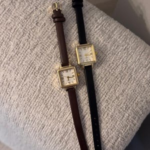 May include: Two square-faced wristwatches with gold-colored cases and Roman numeral hour markers. One watch has a brown leather band, the other a black leather band. Both watches are displayed on a textured, light gray surface.