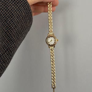 May include: A gold-toned wristwatch with a rectangular face and a gold link band. The watch face is white with black numerals and hands. The watch is held up against a plain background.