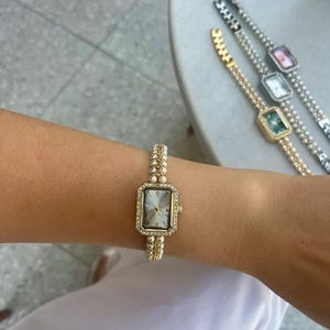 May include: Gold-coloured rectangular watch with a pearl-like strap and a light-coloured face. The watch is decorated with small, sparkling crystals around the face. Several similar watches with different coloured faces are visible in the background.