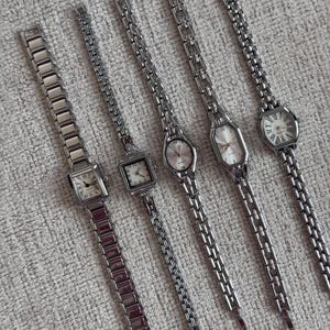 May include: Six silver wristwatches with different face shapes and linked metal straps are shown. The watch faces feature Roman numerals and simple markers. The watches are arranged on a textured, light-coloured surface.