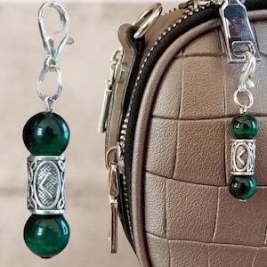 May include: A keychain charm with a silver clasp and chain. The charm features two dark green, spherical beads separated by a silver cylinder with a rune design. The charm is attached to a brown, textured bag.