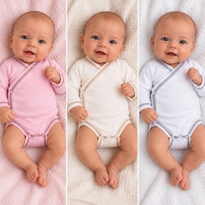 May include: Three infants lying on their backs, each wearing a long-sleeved wrap-style bodysuit. The bodysuits are solid pink, cream, and white, featuring contrasting stitched trim along the crossover neckline, cuffs, and leg openings.