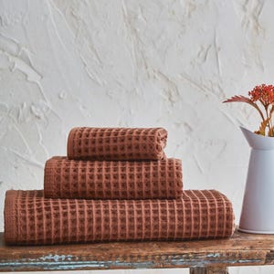 May include: Four stacked, terracotta-colored waffle weave towels on a rustic wooden surface. The towels are neatly folded, showcasing their textured pattern. A white pitcher with orange flowers is in the background, against a textured white wall.