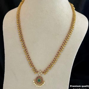May include: A long gold necklace featuring a repeating pattern of small gold beads and embedded red stones along the chain. The central pendant is circular with a green stone at the center, surrounded by red stones, white accents, and small white beads.